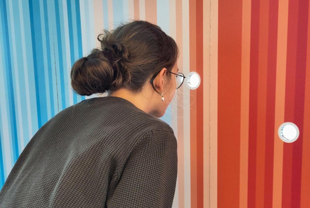 Vrouw kijkt door een rond gaatje in een kleurrijke muur van het klimaatmuseum