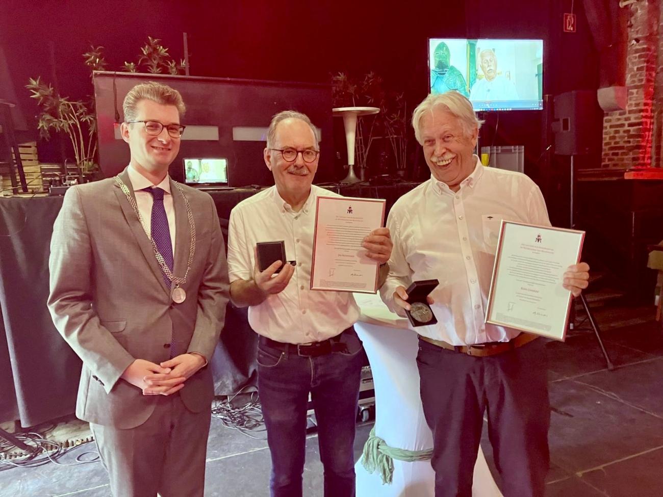Drie mannen poseren tijdens een uitreiking; 2 van hen houden een oorkonde en medaille vast.