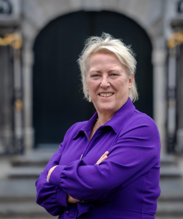 Lachende vrouw met blond haar in een paarse blouse staat met haar armen over elkaar voor het stadhuis
